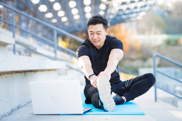 Asian male athlete studies online on the help of a laptop computer, outside the stadium in the park, performs exercises on a remote fitness course - Foto, immagini