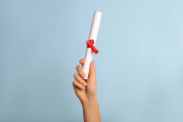 Student holding rolled diploma with red ribbon on light blue background, closeup - Φωτογραφία, εικόνα