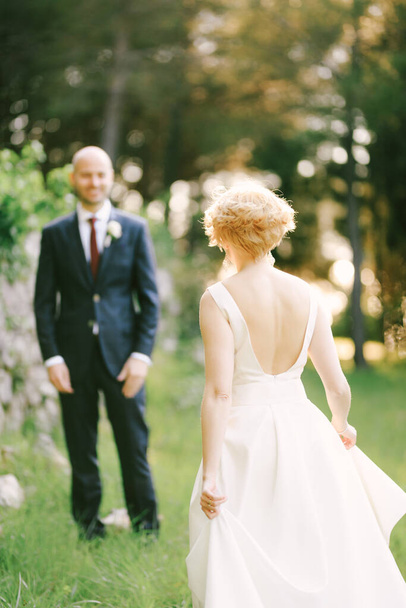 Braut im weißen Kleid geht vor dem Hintergrund grüner Bäume auf lächelnden Bräutigam zu - Foto, Bild