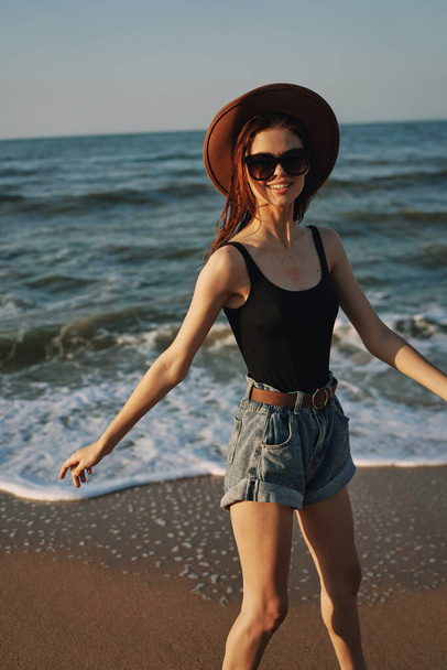 cheerful woman in sunglasses and a hat walks along the ocean - Foto, Imagen