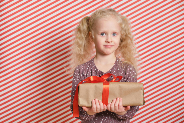 a cute preschooler girl with a gift on a red striped background, a gift with a red ribbon in the hands of a girl with blond hair and two ponytails, boxing day, christmas gift, birthday party - Photo, image