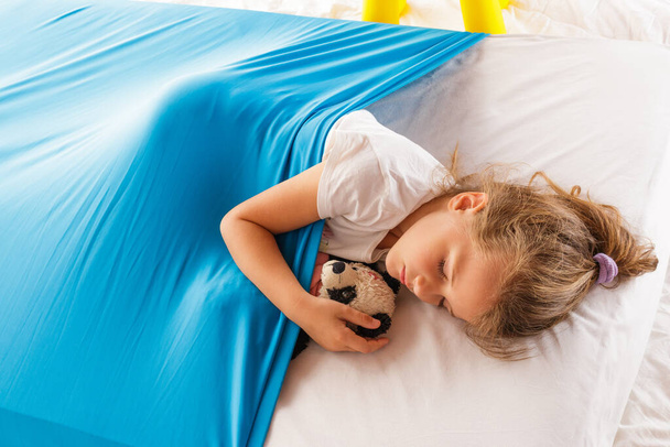 A cute blonde Caucasian girl sleeping on a mattress under a blue cozy blanket - Valokuva, kuva