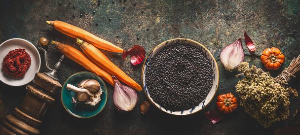 Black lentils with cooking ingredients, vegetables, herbs and spices for tasty vegan meals on dark background. Top view. Healthy vegetarian eating concept. Horizontal banner. Plant based protein - Foto, Imagen