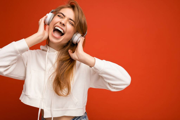 Photo of beautiful attractive happy smiling young blonde woman wearing white hoodie isolated over the colourful background wall with copy space for text wearing white wireless bluetooth earphones - Zdjęcie, obraz