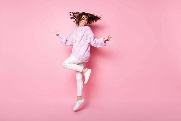 Full length body size view of pretty cheerful wavy-haired girl jumping showing v-sign isolated on pink pastel color background - Foto, afbeelding