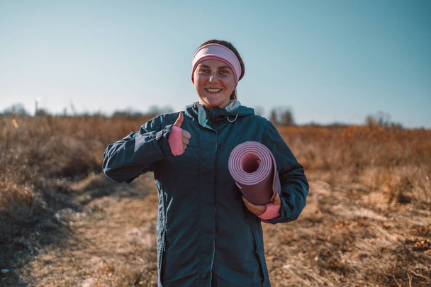 Mujer de yoga muestra como símbolo con la mano sostiene alfombra de ejercicio se ve feliz antes de comenzar un entrenamiento en un parque de otoño. - Foto, Imagen