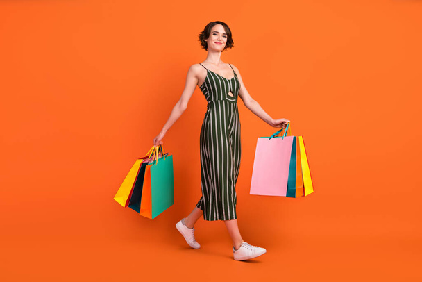 Full length photo of nice brunette millennial lady hold bags go wear overall sneakers isolated on orange background - Fotoğraf, Görsel