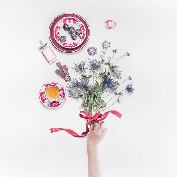 Female hand holding flowers bunch with ribbon on white desktop with cup of tea and women's things. Top view. Flat lay - Photo, image