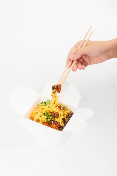 Γυναικείο χέρι που κρατάει ρύζι γουόκ με ξύλινα ξυλάκια, ασιατικό κινέζικο γουόκ που απομονώνεται σε λευκό φόντο. Ένα ανοιχτό κουτί με γουόκ, κοκτέιλ θαλασσινών και λαχανικά. Έννοια παράδοσης γρήγορου φαγητού - Φωτογραφία, εικόνα