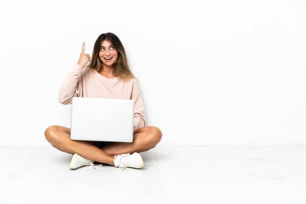 Diz üstü bilgisayarı olan genç bir kadın beyaz arka planda izole bir şekilde oturuyor. Çözümü bulmak için parmağını kaldırıyor. - Fotoğraf, Görsel