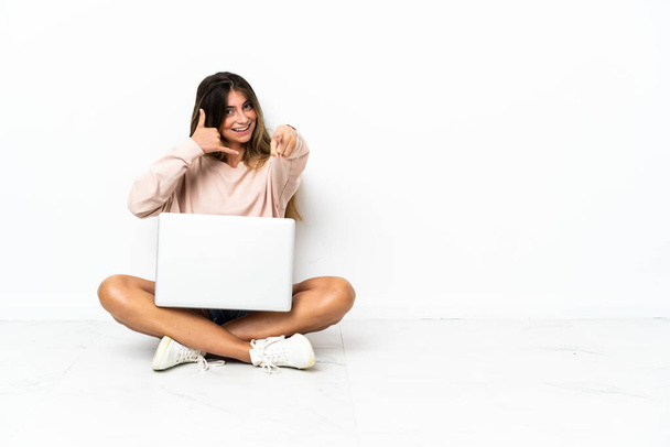 Молода жінка з ноутбуком, що сидить на підлозі ізольовано на білому тлі, роблячи телефонний жест і вказуючи спереду
 - Фото, зображення
