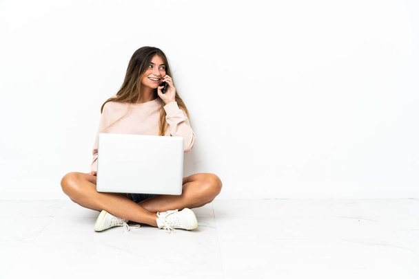 Yerde dizüstü bilgisayarı olan genç bir kadın beyaz arka planda izole bir şekilde oturuyor ve cep telefonuyla sohbet ediyor. - Fotoğraf, Görsel