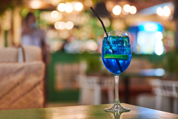 Blue Lagoon cocktail lasi olki, yö kahvila valo bokeh tausta kopioi tilaa. Maukas sininen drinkki kahvilapöydällä. Sininen likööri Curacao maustetulla vodkalla ja limonadilla - Valokuva, kuva