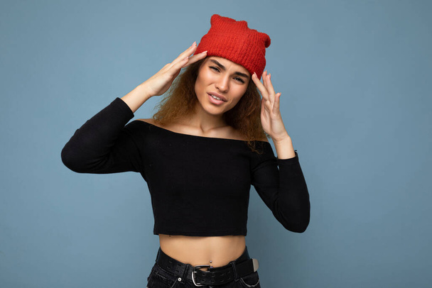 Young emotional dissatisfied pretty brunette woman wavy-haired with sincere emotions wearing black crop top and red hat isolated on blue background with free space and having headache - Fotó, kép