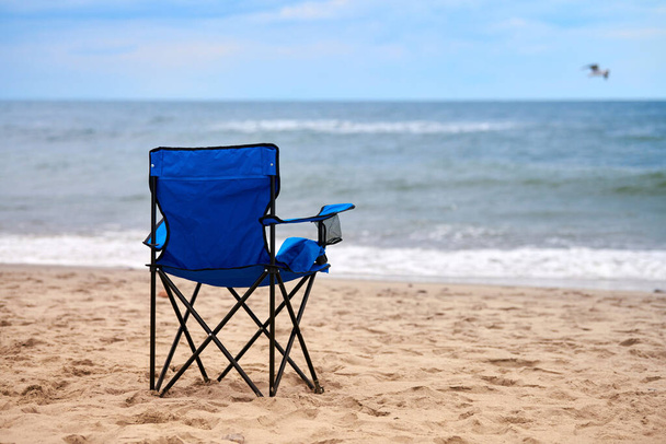 Blue folding chair back on seashore, sea background. Blue chair on sea beach, without people. Beach holiday alone, nobody on Baltic Sea beach, loneliness and hopelessness - Fotoğraf, Görsel