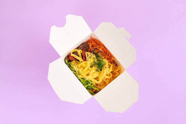 Riisi wok mereneläviä ja vihanneksia valkoinen laatikko eristetty violetti tausta, ylhäältä. Yksi avoin noutorasia riisiwokilla, äyriäiscocktaililla ja vihanneksilla. Aasian ruoka, pikaruoan toimitus käsite - Valokuva, kuva