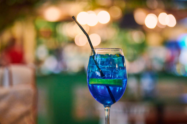 Blue Lagoon cocktail in glas met stro, 's nachts cafe lichte bokeh achtergrond kopieerruimte. Lekker blauw cocktail drankje op cafe tafel. Blauwe likeur Curacao met gearomatiseerde wodka en limonade - Foto, afbeelding