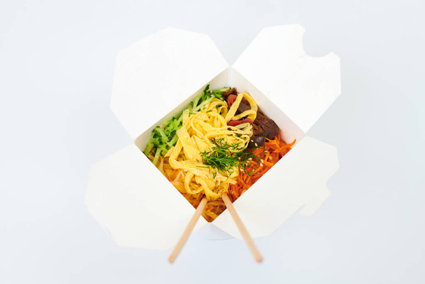 Wok ryżowy z owocami morza i warzywami w białym pudełku i drewniane pałeczki izolowane na białym tle, studio shot. Otwórz pudełko na wynos z wokiem i bambusowymi pałeczkami. Azjatycka żywność, koncepcja fast food - Zdjęcie, obraz