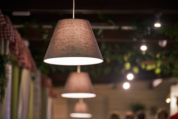 Pendant lamps over tables in city cafe, evening lighting restaurant. Beige fabric lampshades with low dimmed warm light. Modern cozy interior, country style pendant lighting - Photo, Image