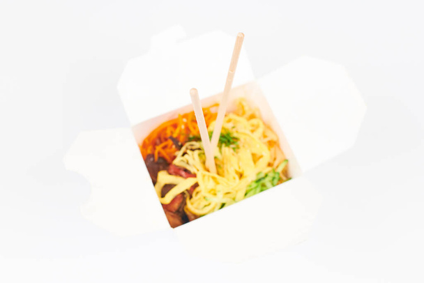 Rice wok with seafood and vegetables in white box and wooden chopsticks isolated on white background, top view, selective focus. Open takeout box with wok and bamboo sticks. Fast food delivery concept - Photo, Image