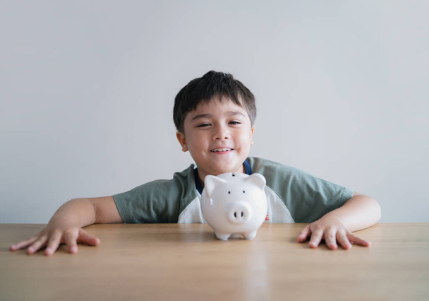 Happy boy siedzi ze świnką bank na stole na białym tle, dziecko z uśmiechniętą twarzą pokazując pieniądze oszczędności.kid Nauka odpowiedzialności finansowej i planowania o oszczędzaniu pieniędzy na przyszłość - Zdjęcie, obraz