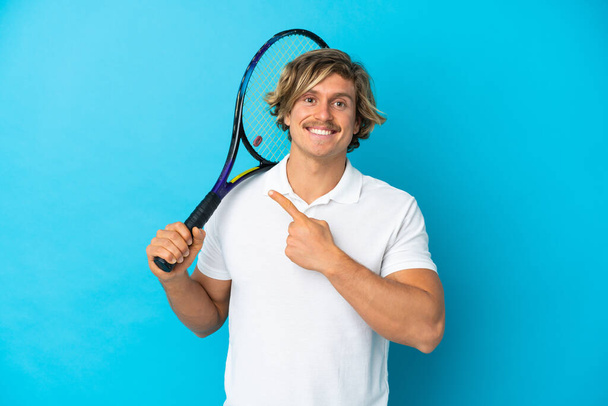 Sarışın tenisçi mavi arka planda izole edilmiş bir adam bir ürünü sunmak için yan tarafı işaret ediyor - Fotoğraf, Görsel