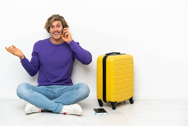 Nuori blondi mies matkalaukku istuu lattialla pitäen keskustelun matkapuhelimen kanssa jonkun - Valokuva, kuva