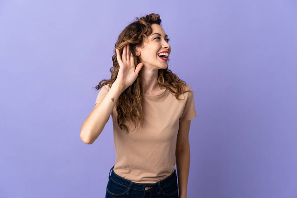 Young caucasian woman isolated on purple background listening to something by putting hand on the ear - Photo, Image