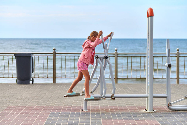 Zelenogradsk, Russie - 07.30.2021 - Jeune fille en vêtements roses à l'aide de la machine d'exercice marcheur d'air, fond marin. Fille engagée dans la forme physique, le jeu de pieds. Exercices sportifs en plein air et air frais marin - Photo, image