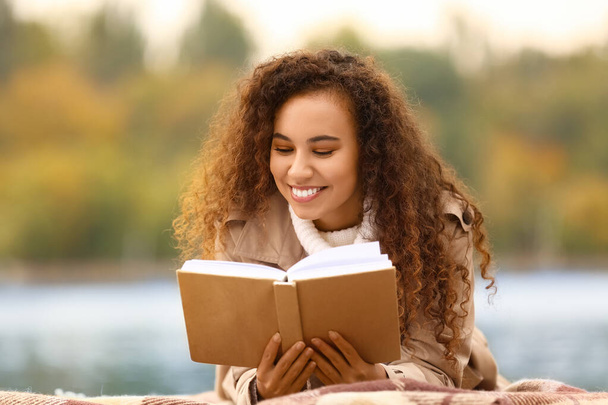 Joven mujer afroamericana leyendo interesante libro sobre cuadros al aire libre - Foto, imagen