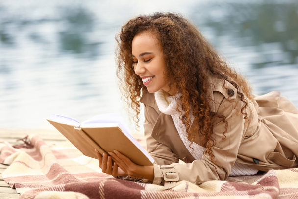 Young African-American woman reading interesting book on plaid near river - Фото, изображение