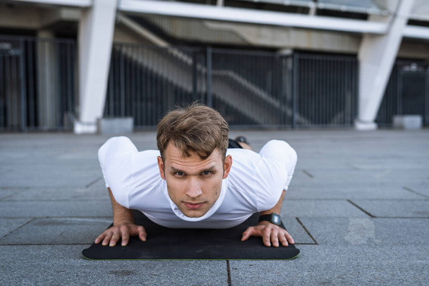Portrait of strong athlete training outdoors, work out on yoga mat, doing push or press ups exercise, look at camera. Confident and concentrated sportsman in sportswear. Physical activity concept - Zdjęcie, obraz