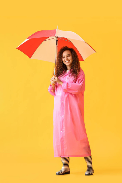 Fiatal afro-amerikai nő rózsaszín esőkabátot és gumicsizmát visel esernyővel sárga alapon - Fotó, kép