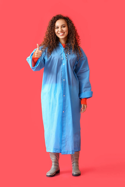 Young African-American woman in blue raincoat showing thumb-up on red background - Foto, Bild