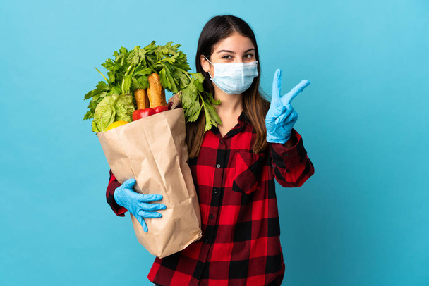 Young caucasian with vegetables and mask isolated on blue background smiling and showing victory sign - Photo, Image
