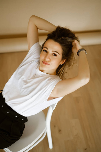 young caucasian woman in black pants and a white T-shirt sitting on a chair, portrait of a brown-eyed brunette with makeup and curly hair, lady in monochrome clothes fixes her hair - 写真・画像