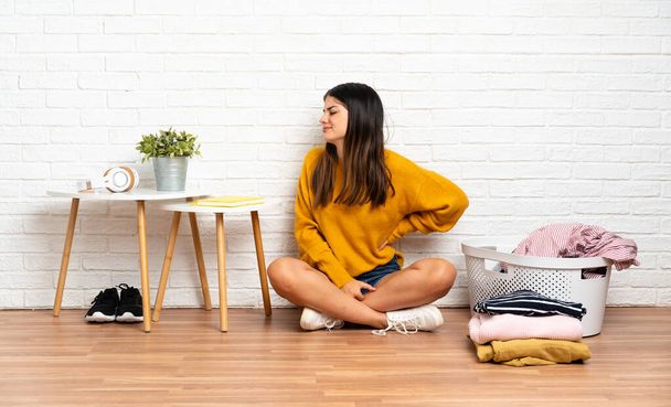 Junge Frau sitzt zu Hause mit Wäschekorb auf dem Boden und leidet unter Rückenschmerzen, weil sie sich bemüht hat - Foto, Bild