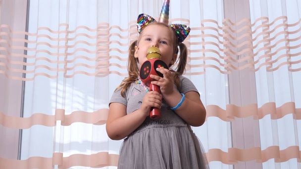 funny cute little girl 4-5 years old, singing into a karaoke microphone, with a unicorn headband, child singing karaoke music, have fun at an event future musician loud voice solo - Photo, Image