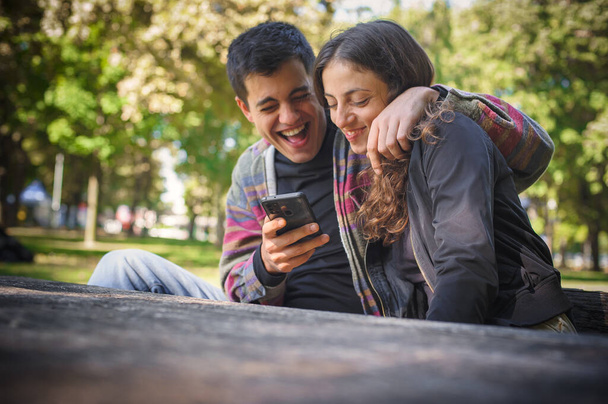 A young smiling couple in love, a boyfriend and a girlfriend, looks at photos together and reads messages on a mobile phone, laugh and enjoy in a public park, outdoors - Fotografie, Obrázek