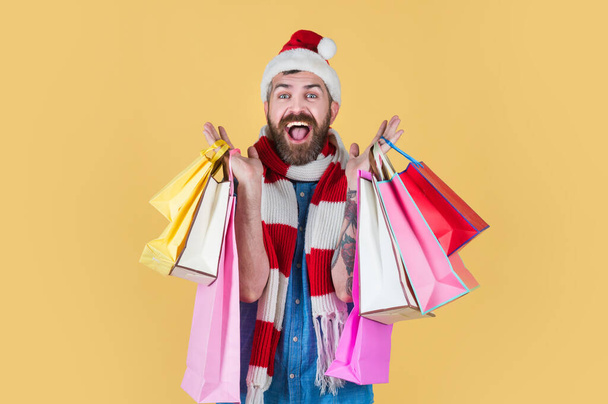 Christmas hipster shopper smile in santa hat and scarf. Man hold shopping bags on yellow background. Man on Christmas - Foto, Imagen