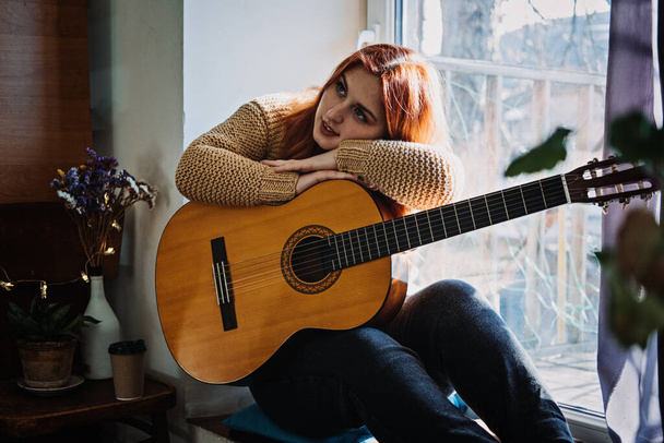 Unaltered candid portrait of young red haired woman in sweater playing acoustic guitar sitting by window at home. Hobbies, indoor home activities for adults in winter, autumn. - Photo, Image
