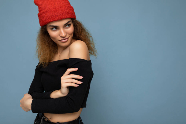 Foto van jonge positieve sexy charmante mooie brunette vrouw krullend met oprechte emoties dragen stijlvolle zwarte gewas top en rode hoed geïsoleerd op blauwe achtergrond met kopieerruimte en op zoek naar de - Foto, afbeelding
