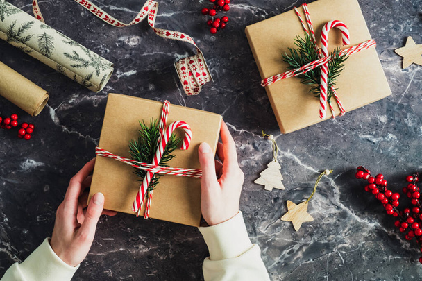 Christmas and New Year holiday preparation concept. Female hand holding gift box with fir tree branch and candy cane over stone table. - Φωτογραφία, εικόνα