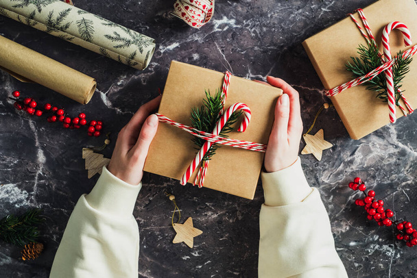 Vrouwelijke handen met kerstcadeau versierd gestreept lint, sparren tak, suikerriet over stenen tafel. Kerstcadeau. - Foto, afbeelding