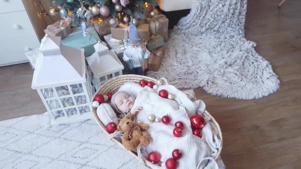 Bebé lindo de cuatro meses que duerme dulcemente en una cesta de mimbre con bolas rojas de Navidad y un oso de peluche en el fondo de un árbol de Navidad y Santa Claus - Metraje, vídeo