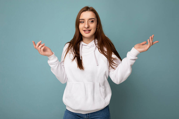 Fotografie mladé pozitivní šťastný krásná brunetka žena s upřímnými emocemi na sobě bílé mikiny izolované přes modré pozadí s kopírovacím prostorem a pokrčení rameny s rukama - Fotografie, Obrázek