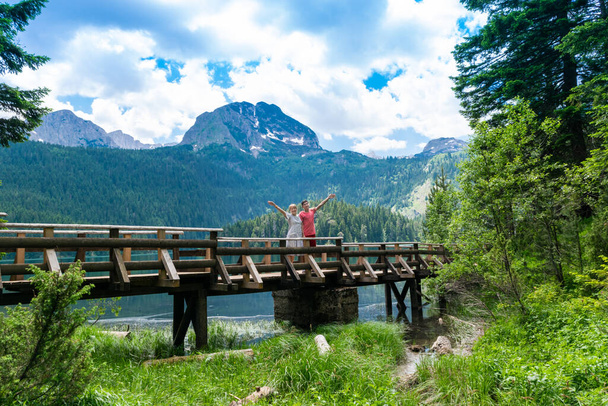 Toeristen op houten voetgangersbrug over de rivier. Meded Peak en Glacial Black Lake achtergrond. Nationaal park Durmitor. Wandelpad in de buurt van het meer is een populaire bestemming voor recreatie en wandelen. Montenegro. - Foto, afbeelding