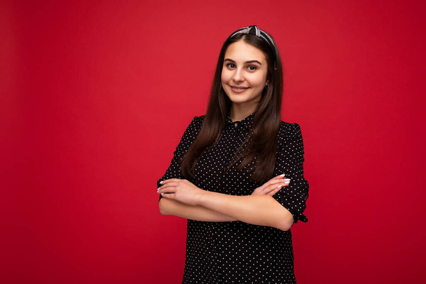 Portrait of young beautiful attractive self-confident positive smiling brunette woman with sincere emotions wearing black polka dot blouse standing with crossed arms isolated on red background with - Photo, Image