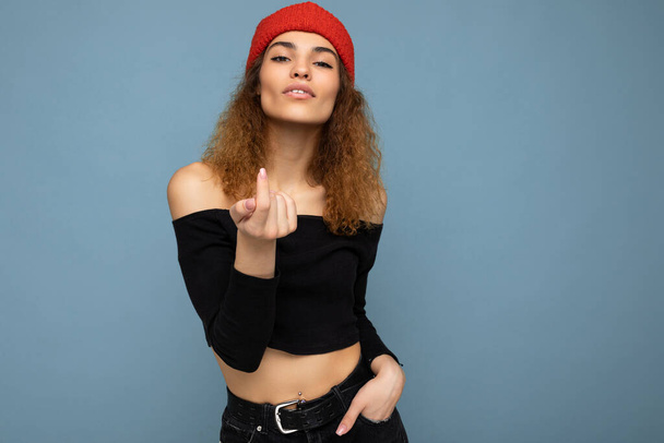 Photo of young positive sexy cute nice brunette woman curly with sincere emotions wearing stylish black crop top and red hat isolated on blue background with copy space and showing beckoning gesture - Photo, Image