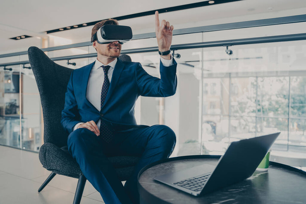 オフィスのロビーに座っている間、 VRヘッドセットゴーグルを身に着けている正式なスーツで手入れの行き届いたCEOの男 - 写真・画像